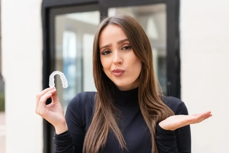 A young woman holding a clear aligner tray with a questioning expression, considering an alternative to traditional dental braces.