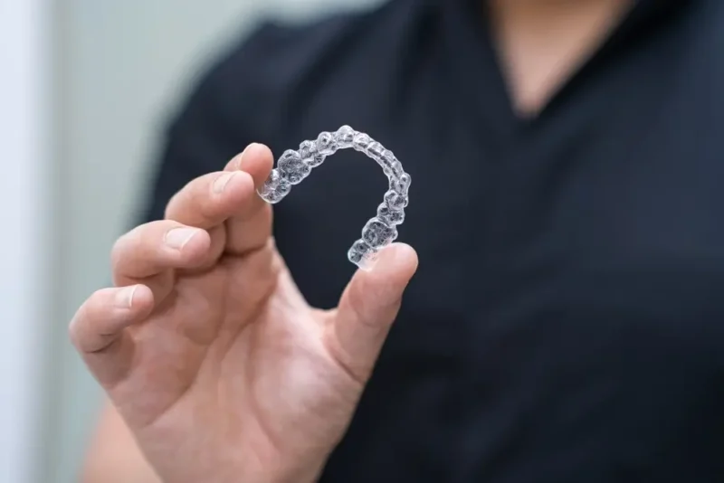 A close-up of a person's hand holding a clear tray of Invisalign braces against a dark background.