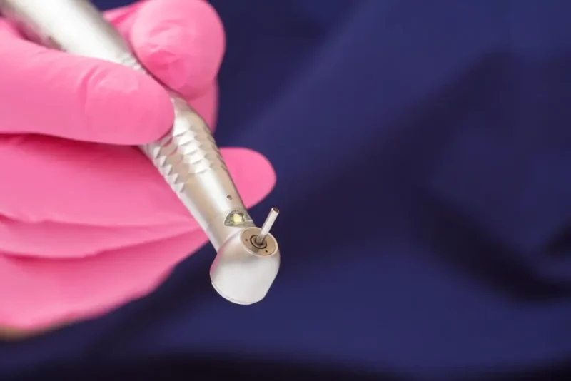 A close-up of a hand in a bright pink glove holding a metallic teeth shaving instrument against a dark blue background.